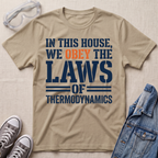 A beige graphic t-shirt laid flat on a textured beige background, styled with safety goggles, gray sneakers, and a blue denim jacket. The t-shirt features the text 'IN THIS HOUSE, WE OBELY THE LAWS OF THERMODYNAMICS' in navy blue and orange.