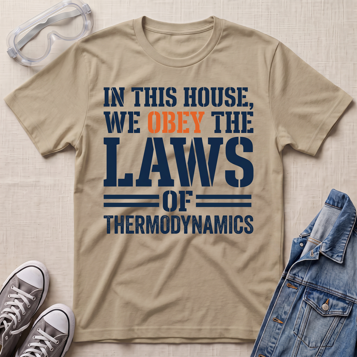 A beige graphic t-shirt laid flat on a textured beige background, styled with safety goggles, gray sneakers, and a blue denim jacket. The t-shirt features the text 'IN THIS HOUSE, WE OBELY THE LAWS OF THERMODYNAMICS' in navy blue and orange.