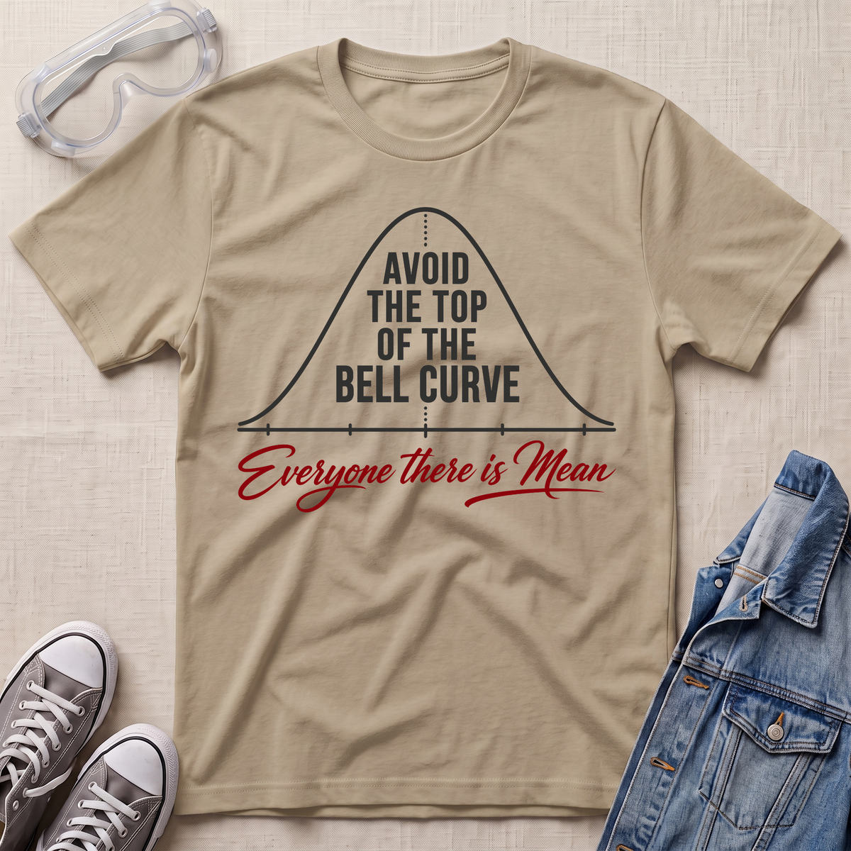 A beige graphic t-shirt laid flat on a textured fabric background, styled with safety goggles, gray sneakers, and a denim jacket. The t-shirt features a black bell curve graphic with the text 'AVOID THE TOP OF THE BELL CURVE' and red script text reading 'Everyone there is Mean'.