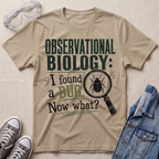 A beige graphic t-shirt featuring the text 'OBSERVATIONAL BIOLOGY: I found a BUG. Now what?' in dark green and black lettering, accompanied by a magnifying glass and beetle illustration.