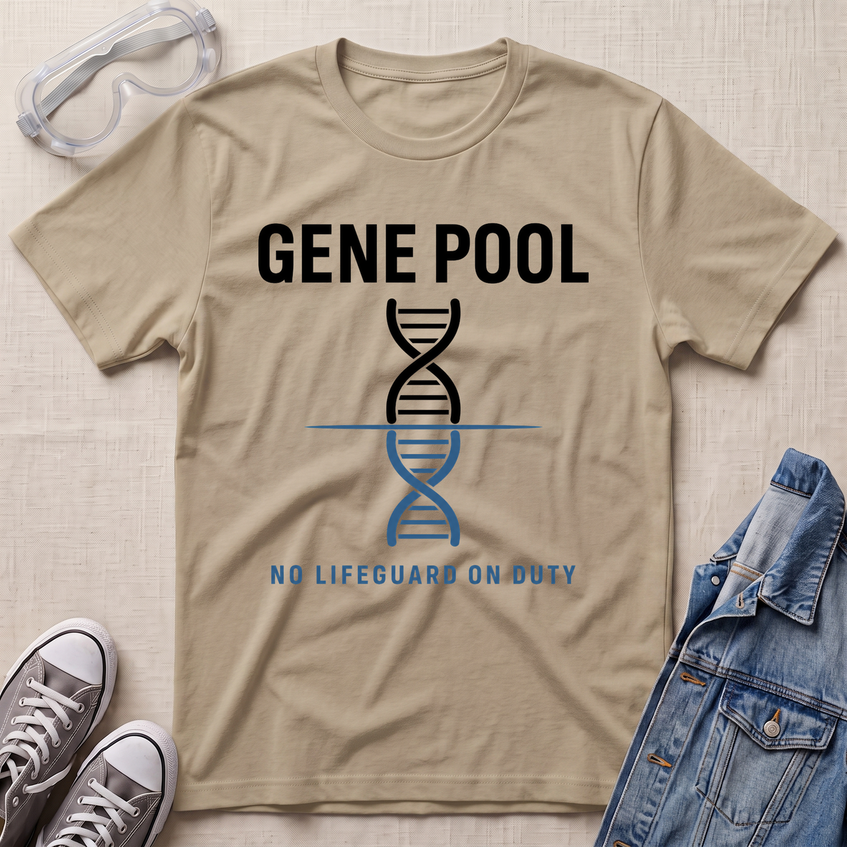 A beige short-sleeve t-shirt laid flat on a textured light background, featuring a black and blue DNA double helix graphic with the text 'GENE POOL' and 'NO LIFEGUARD ON DUTY'. Goggles, gray sneakers, and a blue denim jacket are styled around the shirt.