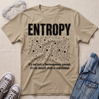 A beige graphic t-shirt laid flat on a beige fabric background, styled with safety goggles, gray sneakers, and a denim jacket. The t-shirt features a white distressed graphic of the word 'ENTROPY' above a entropy pattern, with the text 'It's not just a thermodynamic concept, it's my default state of organization' below.