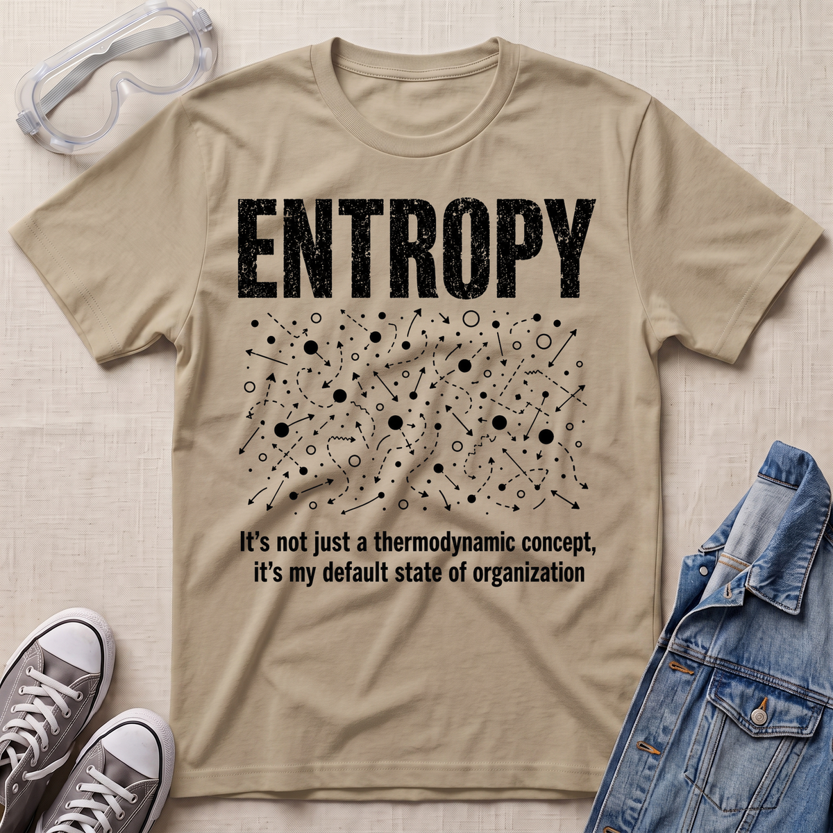 A beige graphic t-shirt laid flat on a beige fabric background, styled with safety goggles, gray sneakers, and a denim jacket. The t-shirt features a white distressed graphic of the word 'ENTROPY' above a entropy pattern, with the text 'It's not just a thermodynamic concept, it's my default state of organization' below.