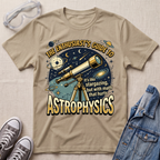 A sand beige graphic T-shirt laid flat on a light fabric surface, styled with clear safety goggles, gray sneakers, and a blue denim jacket. The shirt features a large space-themed print with a telescope, planets, stars, and the text 'THE ENTHUSIAST'S GUIDE TO ASTROPHYSICS' and 'it's like stargazing, but with math that hurts.