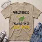 A beige graphic t-shirt laid flat on a textured fabric background, surrounded by styling props including safety goggles, gray sneakers, and a denim jacket. The t-shirt features a green leaf graphic and the text 'PHOTOSYNTHESIS I'M A BIG FAN OF LIGHT WORK' in black and yellow.