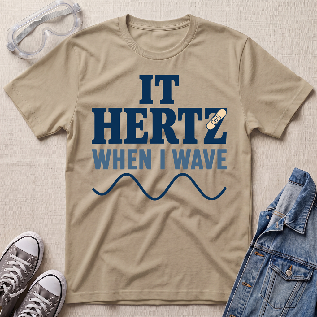 A beige graphic t-shirt laid flat on a textured light beige background, styled with safety goggles, gray sneakers, and a denim jacket. The t-shirt features a dark blue and light blue graphic that reads 'IT HERTZ WHEN I WAVE' with a wavy line and a band-aid on an EKG line.
