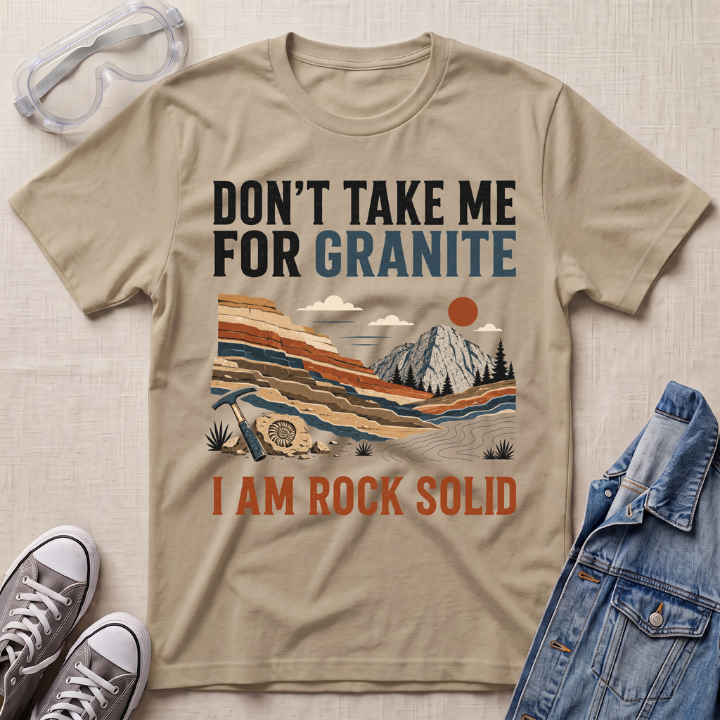 A beige graphic t-shirt laid flat on a textured beige fabric background, surrounded by styling props including safety goggles, gray sneakers, and a blue denim jacket. The t-shirt features a retro landscape illustration of a mountain and layered rock formations, with the text 'DON'T TAKE ME FOR GRANITE' above and 'I AM ROCK SOLID' below.