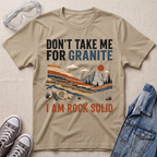 A beige graphic t-shirt laid flat on a textured beige fabric background, surrounded by styling props including safety goggles, gray sneakers, and a blue denim jacket. The t-shirt features a retro landscape illustration of a mountain and layered rock formations, with the text 'DON'T TAKE ME FOR GRANITE' above and 'I AM ROCK SOLID' below.