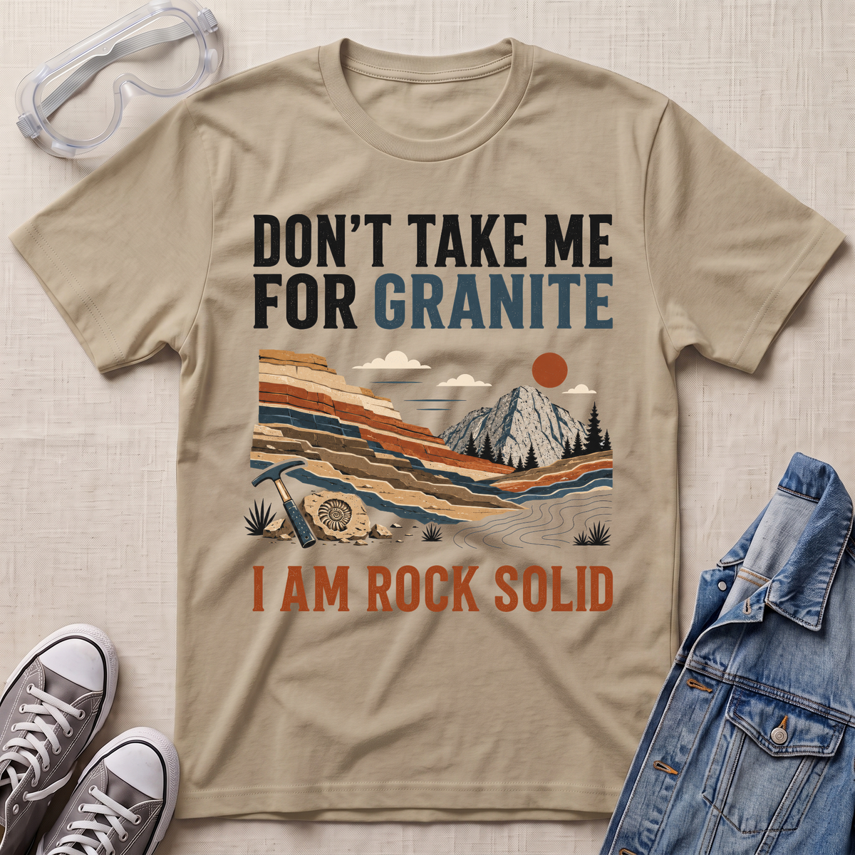 A beige graphic t-shirt laid flat on a textured beige fabric background, surrounded by styling props including safety goggles, gray sneakers, and a blue denim jacket. The t-shirt features a retro landscape illustration of a mountain and layered rock formations, with the text 'DON'T TAKE ME FOR GRANITE' above and 'I AM ROCK SOLID' below.