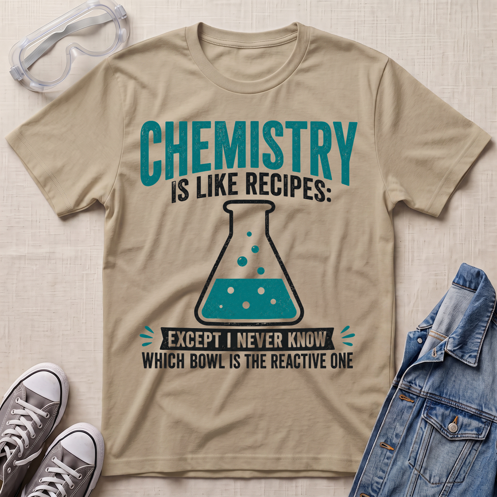 A beige graphic t-shirt laid flat on a beige fabric background, styled with safety goggles, gray sneakers, and a denim jacket. The t-shirt features a teal and black graphic that reads 'CHEMISTRY IS LIKE RECIPES: EXCEPT I NEVER KNOW WHICH BOWL IS THE REACTIVE ONE' with an illustration of a beaker.