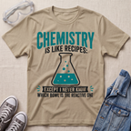 A beige graphic t-shirt laid flat on a beige fabric background, styled with safety goggles, gray sneakers, and a denim jacket. The t-shirt features a teal and black graphic that reads 'CHEMISTRY IS LIKE RECIPES: EXCEPT I NEVER KNOW WHICH BOWL IS THE REACTIVE ONE' with an illustration of a beaker.
