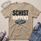 A beige graphic t-shirt laid flat on a textured beige background, surrounded by styling props including safety goggles, gray sneakers, and a blue denim jacket. The t-shirt features the text 'SCHIST HAPPENS' in bold black and cream lettering above an illustration of a dark gray schist rock.