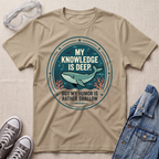 A beige graphic t-shirt laid flat on a textured beige fabric background, styled with safety goggles, gray sneakers, and a denim jacket. The t-shirt features a circular graphic with a teal whale and the text 'MY KNOWLEDGE IS DEEP BUT MY HUMOR IS RATHER SHALLOW'.