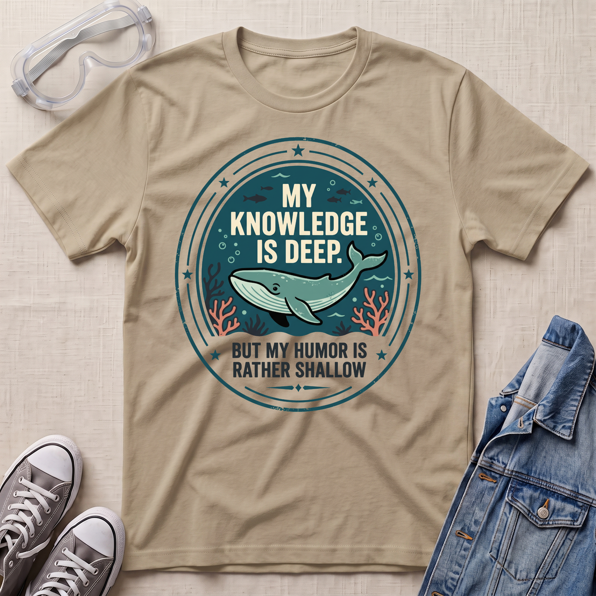 A beige graphic t-shirt laid flat on a textured beige fabric background, styled with safety goggles, gray sneakers, and a denim jacket. The t-shirt features a circular graphic with a teal whale and the text 'MY KNOWLEDGE IS DEEP BUT MY HUMOR IS RATHER SHALLOW'.