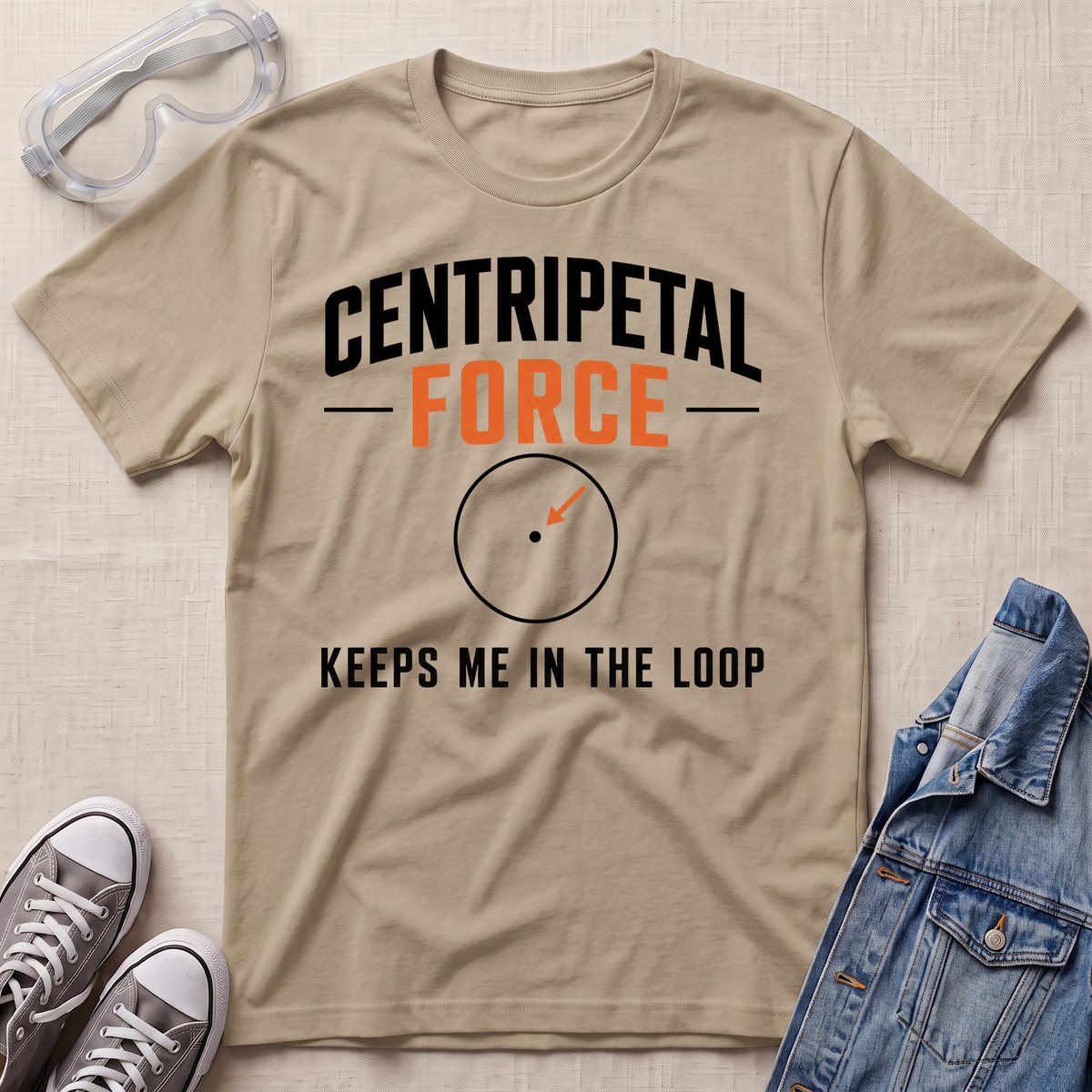 A beige graphic t-shirt laid flat on a textured light background, styled with safety goggles, gray sneakers, and a denim jacket. The t-shirt features the text 'CENTRIPETAL FORCE' in black and orange, with an arrow graphic below.