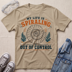 A beige graphic t-shirt laid flat on a textured fabric background, surrounded by styling props including clear safety goggles, gray sneakers, and a blue denim jacket. The t-shirt features a retro-style graphic with a spiral ammonite shell and the text 'MY LIFE IS SPIRALING OUT OF CONTROL' in orange and navy blue.