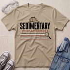 A beige graphic t-shirt featuring a detective themed 'Sedimentary My Dear Rockson' print, styled in a flat-lay with a denim jacket, sneakers, and safety goggles on a textured beige background.