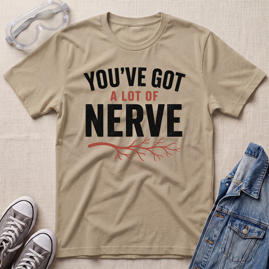 A beige graphic t-shirt with the text 'YOU'VE GOT A LOT OF NERVE' in black and red, styled in a flat lay with safety goggles, gray sneakers, and a denim jacket on a light beige textured background.