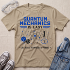 A beige graphic t-shirt featuring a blue and black 'Quantum Mechanics is Easy' design, styled in a flat-lay with safety goggles, sneakers, and a denim jacket on a textured beige background.