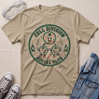 A beige graphic t-shirt laid flat on a textured fabric background, styled with safety goggles, gray sneakers, and a denim jacket. The t-shirt features a circular retro-style graphic reading 'CELL DIVISION SOCIAL CLUB' with cartoon cells and lab-themed illustrations.