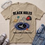 A sand beige graphic t-shirt laid flat on a textured fabric background, styled with safety goggles, sneakers, and a denim jacket. The t-shirt features a cartoon illustration of a vacuum cleaner falling into a black hole surrounded by planets, with the text 'BLACK HOLES' and 'NATURE'S ULTIMATE CLEANING SERVICE'.