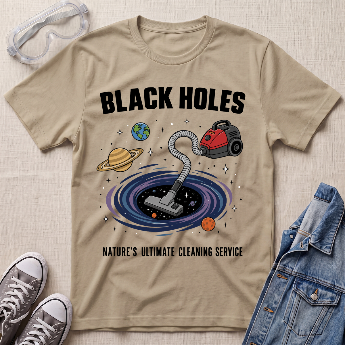 A sand beige graphic t-shirt laid flat on a textured fabric background, styled with safety goggles, sneakers, and a denim jacket. The t-shirt features a cartoon illustration of a vacuum cleaner falling into a black hole surrounded by planets, with the text 'BLACK HOLES' and 'NATURE'S ULTIMATE CLEANING SERVICE'.