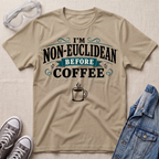 A beige graphic t-shirt laid flat on a textured light beige background, styled with safety goggles, gray sneakers, and a blue denim jacket. The t-shirt features the text 'I'M NON-EUCLIDEAN BEFORE COFFEE' in black and teal lettering with a coffee cup illustration below.