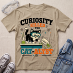 A beige graphic T-shirt laid flat on a light fabric surface, styled with safety goggles, gray sneakers, and a denim jacket. The shirt features a cartoon black-and-white cat wearing blue goggles and leaning over a counter with green substance spilling out, surrounded by text reading “CURIOSITY KILLED THE CAT-ALYST.