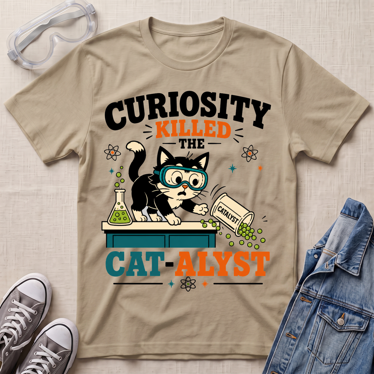 A beige graphic T-shirt laid flat on a light fabric surface, styled with safety goggles, gray sneakers, and a denim jacket. The shirt features a cartoon black-and-white cat wearing blue goggles and leaning over a counter with green substance spilling out, surrounded by text reading “CURIOSITY KILLED THE CAT-ALYST.