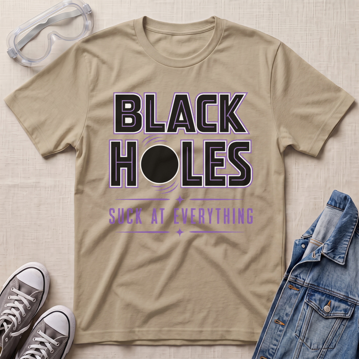 A sand beige graphic t-shirt laid flat on a textured beige background, styled with safety goggles, gray sneakers, and a denim jacket. The t-shirt features a graphic that reads 'BLACK HOLES SUCK AT EVERYTHING' in black and purple text with a black circle in the center.