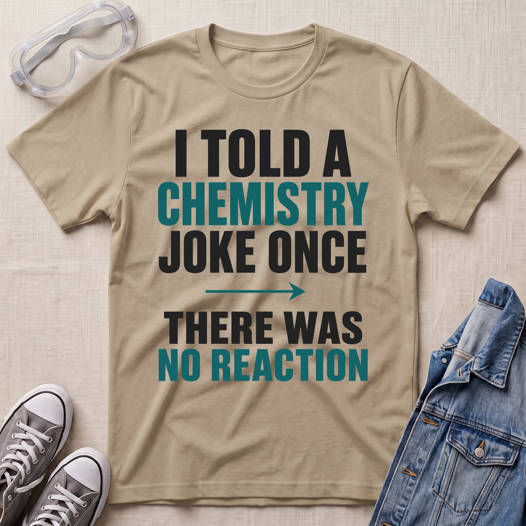A beige graphic t-shirt with the text 'I TOLD A CHEMISTRY JOKE ONCE THERE WAS NO REACTION' in black and teal lettering, styled in a flat-lay with safety goggles, gray sneakers, and a denim jacket on a textured beige background.