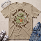 A beige graphic t-shirt laid flat on a textured light beige surface, surrounded by styled props including clear safety goggles, gray sneakers, and a blue denim jacket. The t-shirt features a circular retro-style graphic of a green creature wearing goggles and holding a mug, with the text 'LABORATORY SURVIVAL CLUB' and 'UNDER PRESSURE, BUT ADAPTING'.