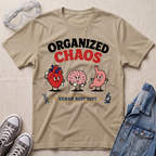 A beige graphic t-shirt laid flat on a textured fabric background, styled with safety goggles, gray sneakers, and a denim jacket. The t-shirt features a cartoon graphic of a red heart, a pink brain, and a pink stomach with the text 'ORGANIZED CHAOS' and 'HUMAN BODY DEPT.