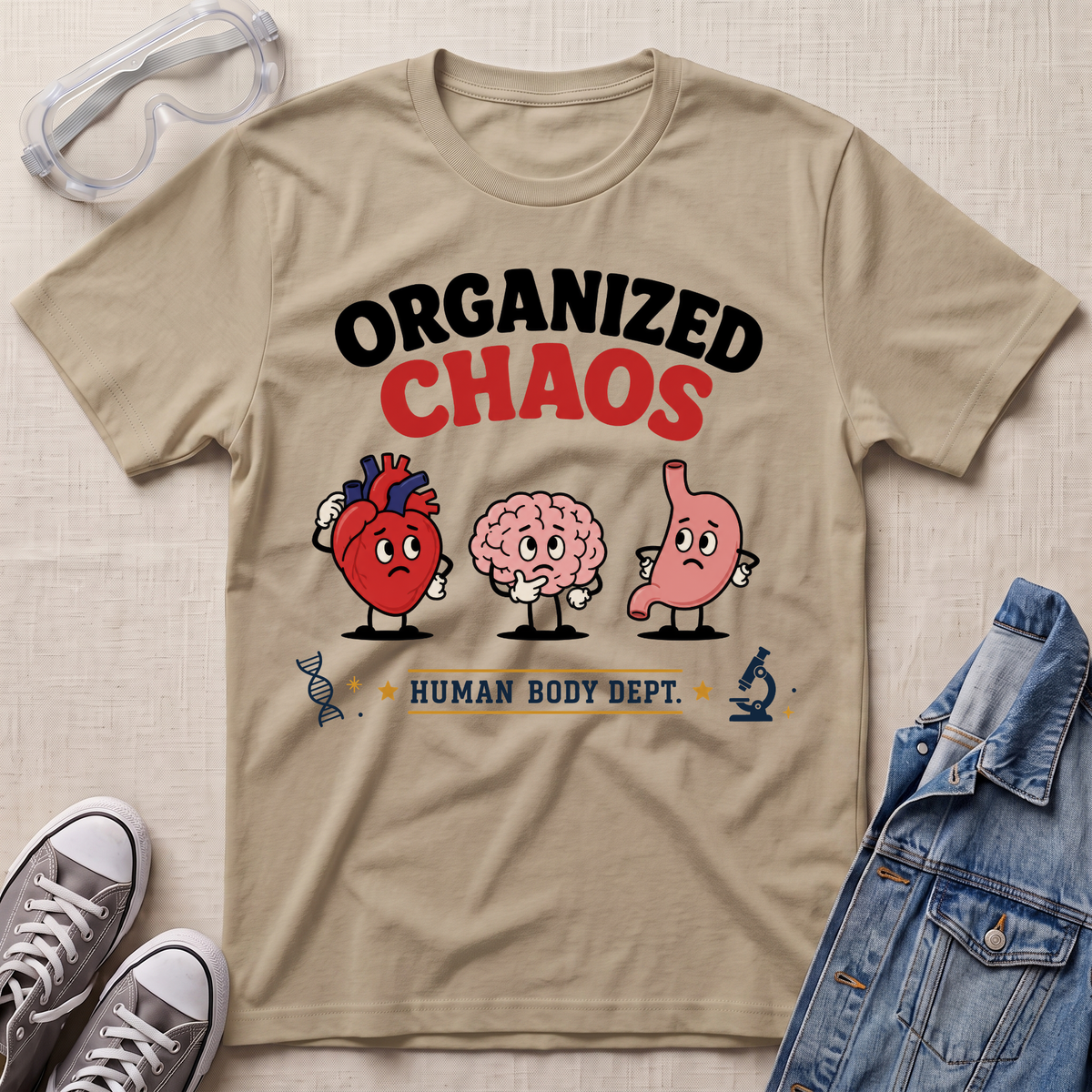 A beige graphic t-shirt laid flat on a textured fabric background, styled with safety goggles, gray sneakers, and a denim jacket. The t-shirt features a cartoon graphic of a red heart, a pink brain, and a pink stomach with the text 'ORGANIZED CHAOS' and 'HUMAN BODY DEPT.