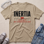 A beige graphic t-shirt laid flat on a textured light beige background, surrounded by styling props including clear safety goggles, gray sneakers, and a blue denim jacket. The t-shirt features the word 'INERTIA' in bold black letters, with a small red block and red text underneath reading 'I'D EXPLAIN IT, BUT I'D RATHER STAY AT REST'.