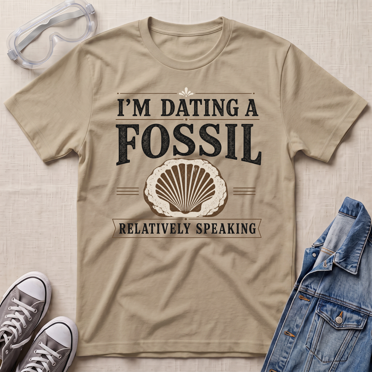 A beige graphic t-shirt laid flat on a textured fabric background, surrounded by styling props including safety goggles, gray sneakers, and a denim jacket. The t-shirt features a distressed black and beige graphic that reads 'I'M DATING A FOSSIL RELATIVELY SPEAKING' with a seashell illustration.