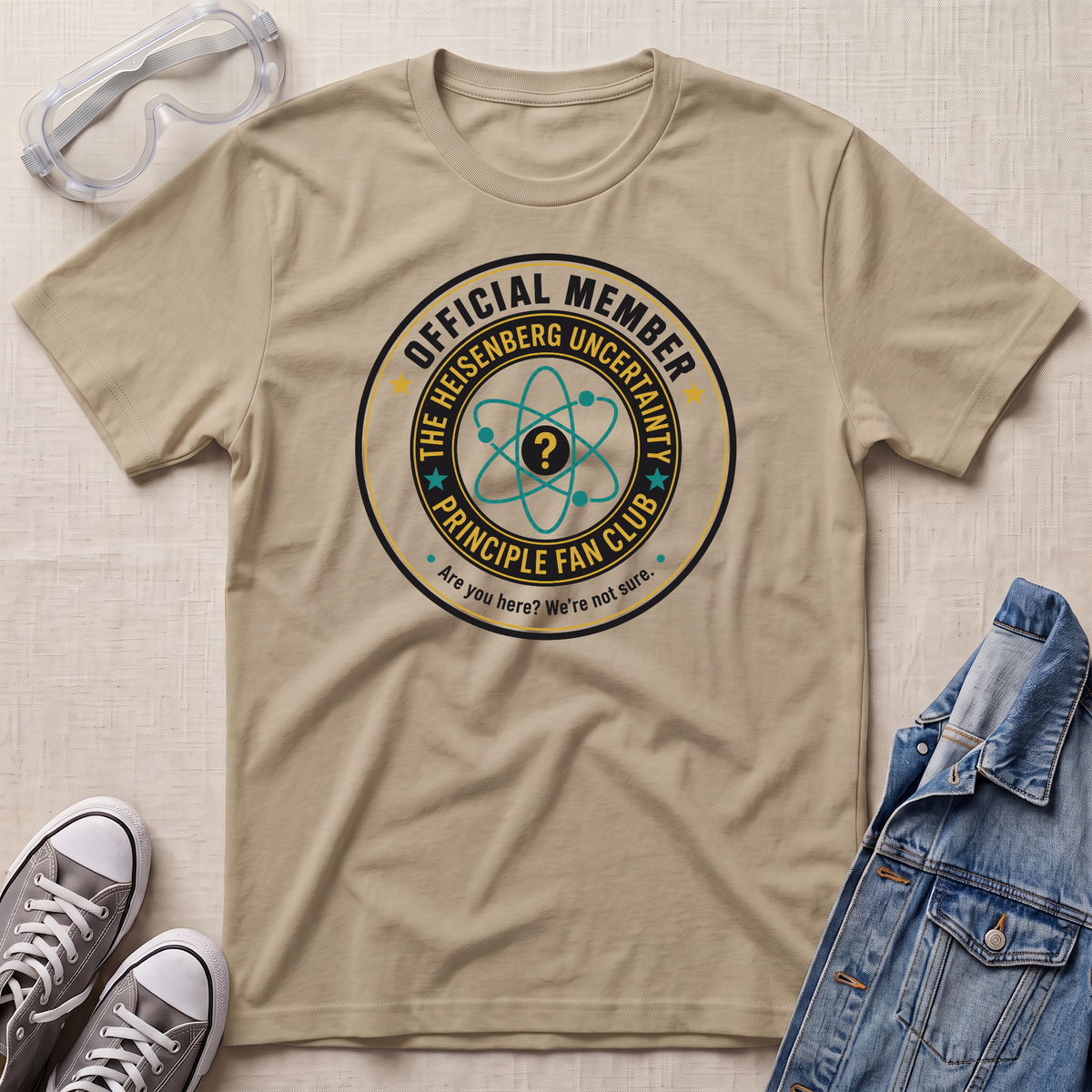 A beige graphic t-shirt laid flat on a textured light beige surface, styled with safety goggles, gray sneakers, and a blue denim jacket. The t-shirt features a circular graphic that reads 'OFFICIAL MEMBER THE HEISENBERG UNCERTAINTY PRINCIPLE FAN CLUB' with an atom symbol.