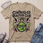 A beige graphic t-shirt featuring a green beaker with sunglasses and the text 'CONTROLLED REACTION CLUB' in black, styled in a flat lay with safety goggles, sneakers, and a denim jacket on a textured beige background.