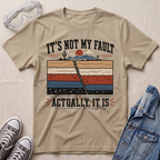 A beige graphic t-shirt laid flat on a textured fabric background, styled with safety glasses, gray sneakers, and a blue denim jacket. The t-shirt features a retro-style desert landscape graphic with a cracked earth design and the text 'IT'S NOT MY FAULT ACTUALLY, IT IS'.