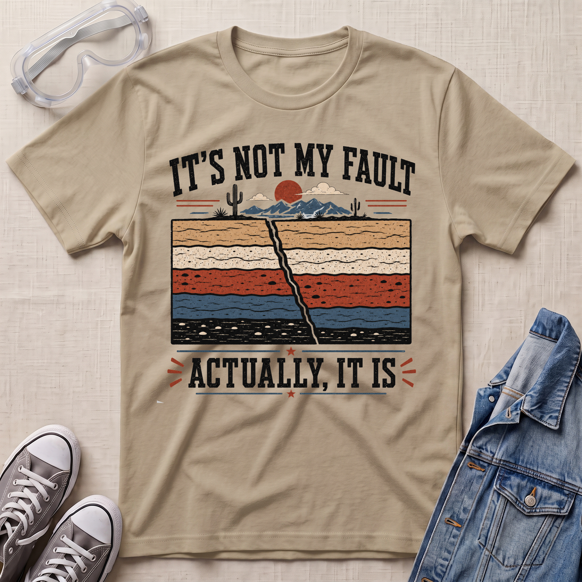 A beige graphic t-shirt laid flat on a textured fabric background, styled with safety glasses, gray sneakers, and a blue denim jacket. The t-shirt features a retro-style desert landscape graphic with a cracked earth design and the text 'IT'S NOT MY FAULT ACTUALLY, IT IS'.