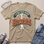 A beige graphic t-shirt laid flat on a light wood surface, featuring a bone graphic and the text 'I FIND THIS SHIRT HUMERUS' in orange and green. The shirt is styled with safety glasses, gray sneakers, and a denim jacket.