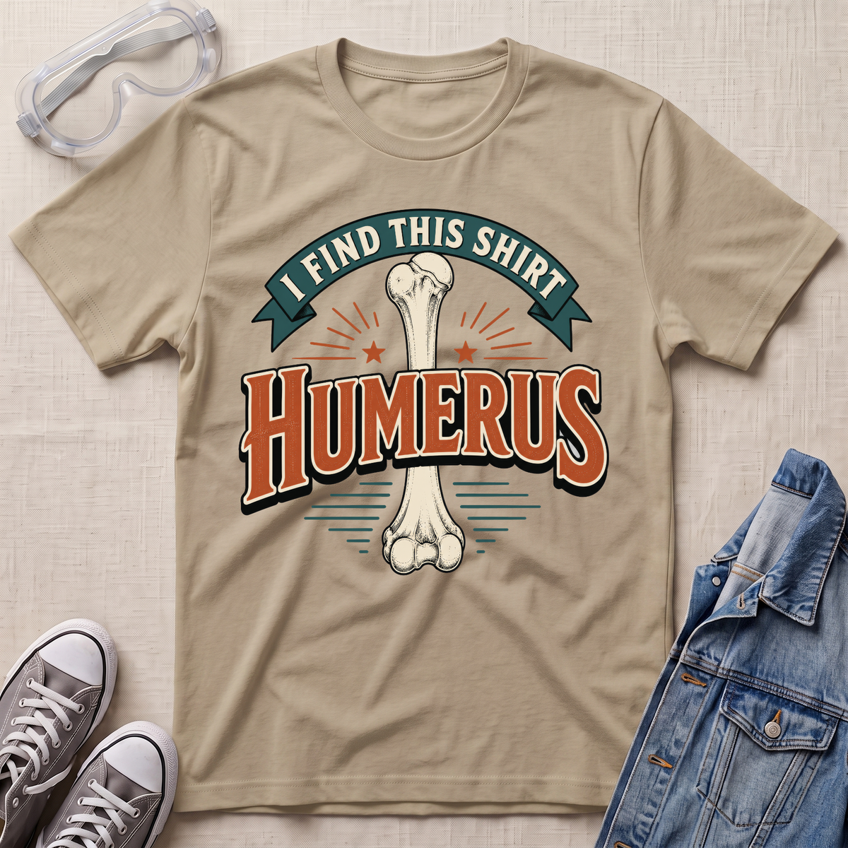 A beige graphic t-shirt laid flat on a light wood surface, featuring a bone graphic and the text 'I FIND THIS SHIRT HUMERUS' in orange and green. The shirt is styled with safety glasses, gray sneakers, and a denim jacket.