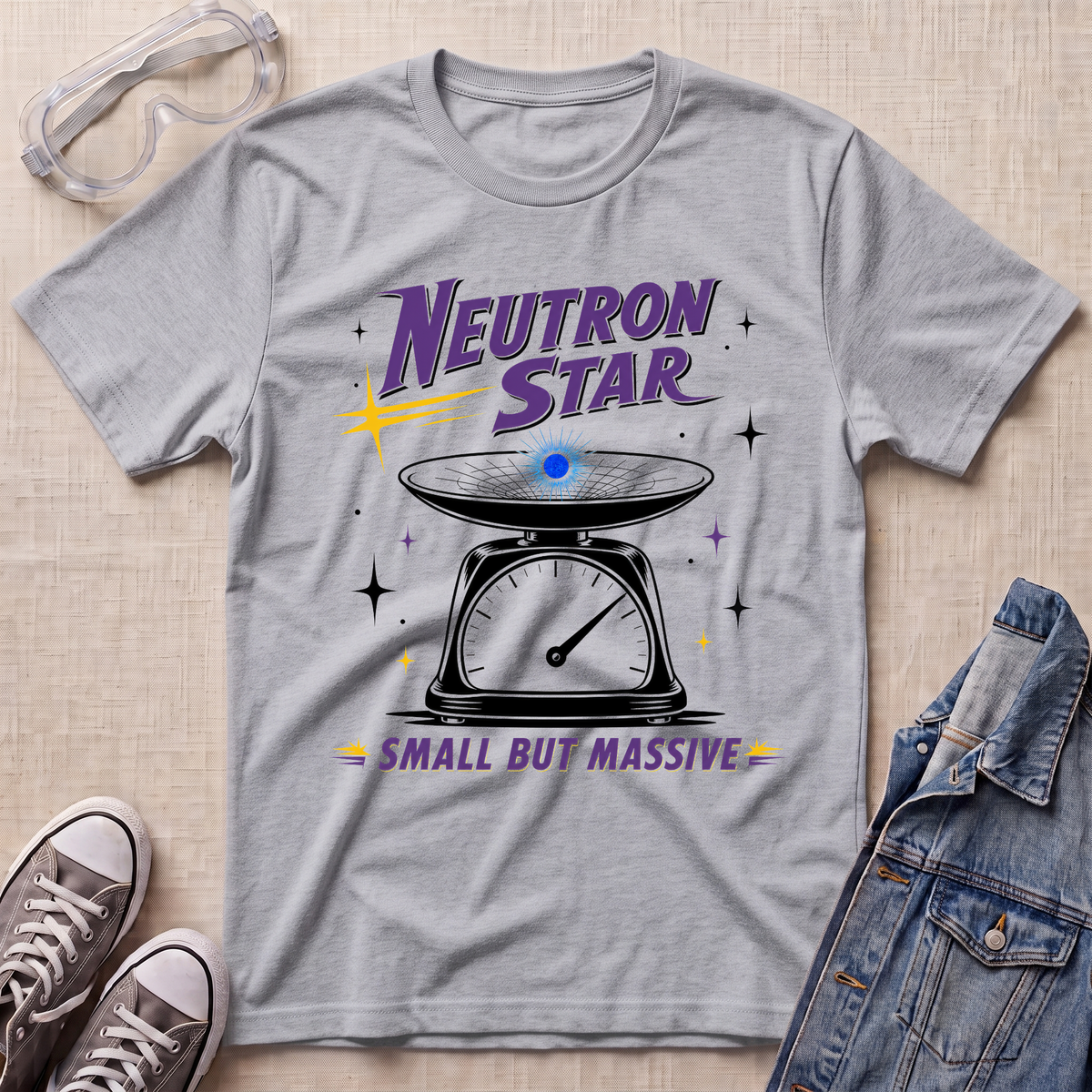 A heather sport gray graphic t-shirt featuring a retro-style 'Neutron Star' design with a scale illustration and the phrase 'Small But Massive'. The t-shirt is styled in a flat-lay arrangement on a beige textured background, surrounded by props including safety goggles, sneakers, and a denim jacket.