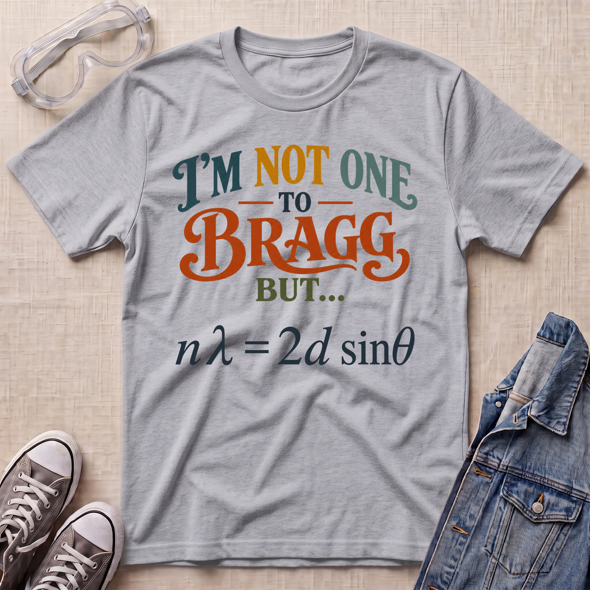 A heather gray graphic t-shirt laid flat on a beige textured background, styled with safety goggles, sneakers, and a denim jacket. The t-shirt features a humorous physics joke printed in orange, teal, and dark gray text.