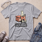 A heather gray short-sleeve t-shirt laid flat on a beige textured surface, featuring a cartoon graphic of a lab flask character sitting next to a red-orange box with the text 'Chemically Exhausted'. Surrounding props include safety goggles, gray sneakers, and a denim jacket.