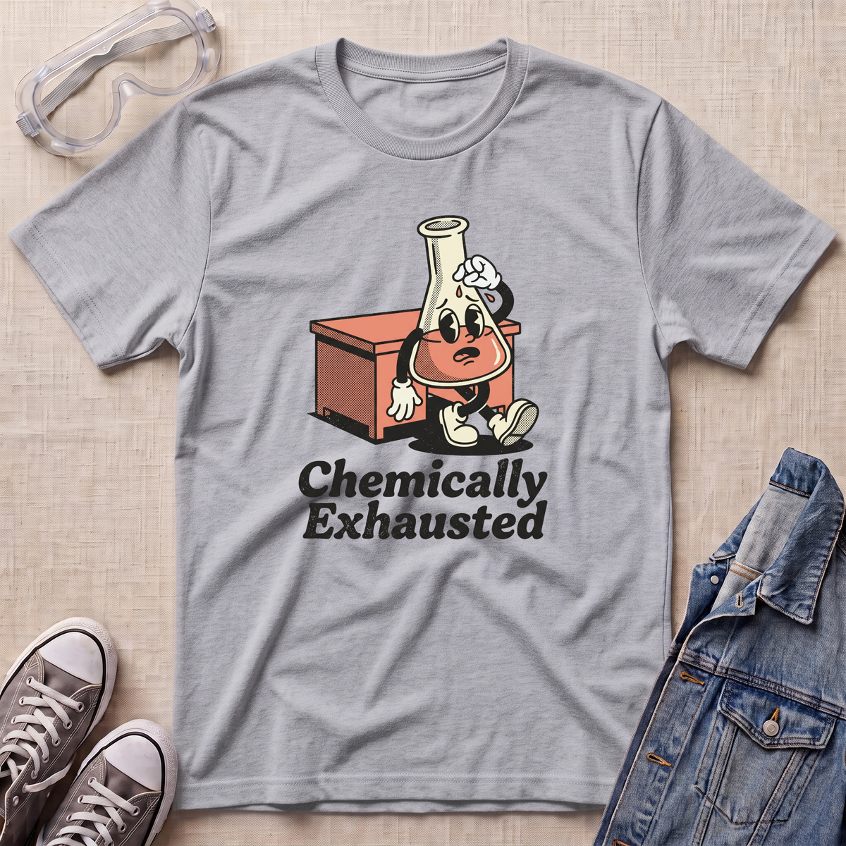 A heather gray short-sleeve t-shirt laid flat on a beige textured surface, featuring a cartoon graphic of a lab flask character sitting next to a red-orange box with the text 'Chemically Exhausted'. Surrounding props include safety goggles, gray sneakers, and a denim jacket.
