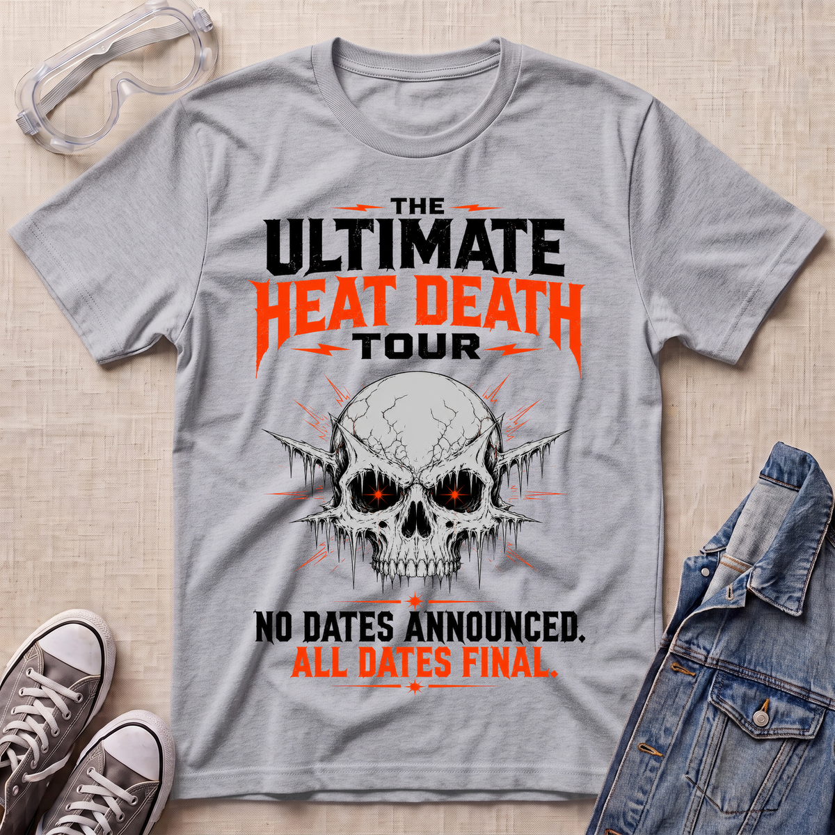 A heather gray graphic t-shirt laid flat on a beige textured surface, styled with safety glasses, gray sneakers, and a denim jacket. The t-shirt features a skull graphic with glowing red eyes and bold text reading 'THE ULTIMATE HEAT DEATH TOUR' and 'NO DATES ANNOUNCED.