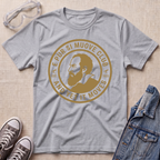A heather sport gray short-sleeve graphic T-shirt laid flat on a light beige textured surface, surrounded by styling props including clear safety goggles, gray sneakers, and a blue denim jacket. The shirt features a large circular tan/gold print with a portrait and the text 'E PUR SI MUOVE CLUB AND YET IT MOVES'.
