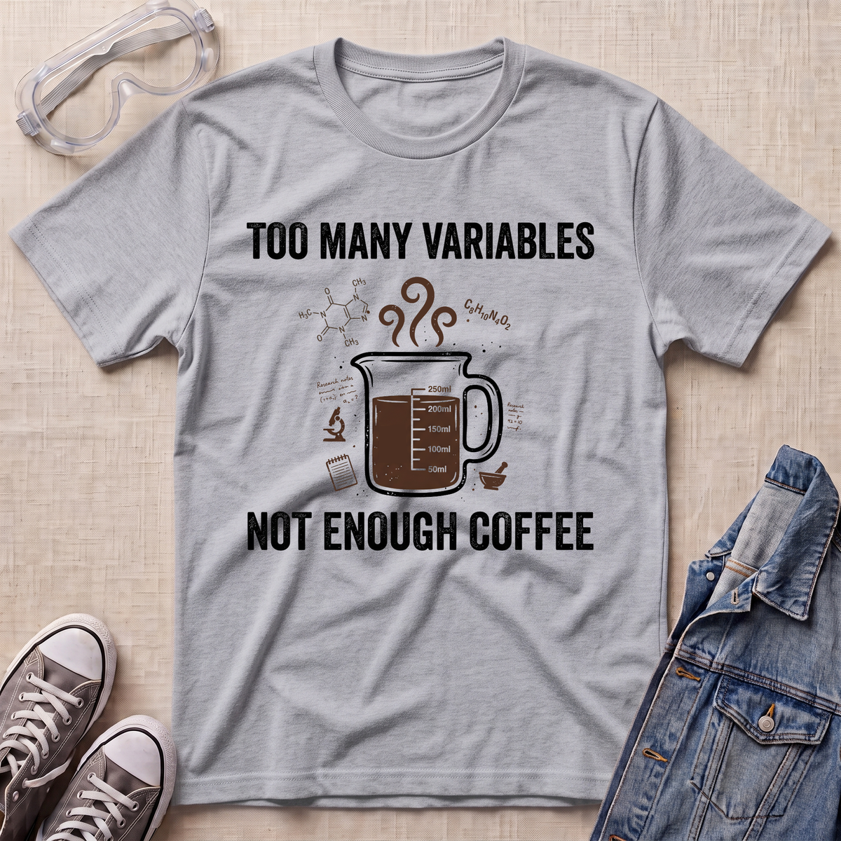 A heather gray graphic t-shirt laid flat on a beige textured background, styled with safety goggles, sneakers, and a denim jacket. The t-shirt features a brown coffee mug graphic with steam and the text 'TOO MANY VARIABLES NOT ENOUGH COFFEE'.