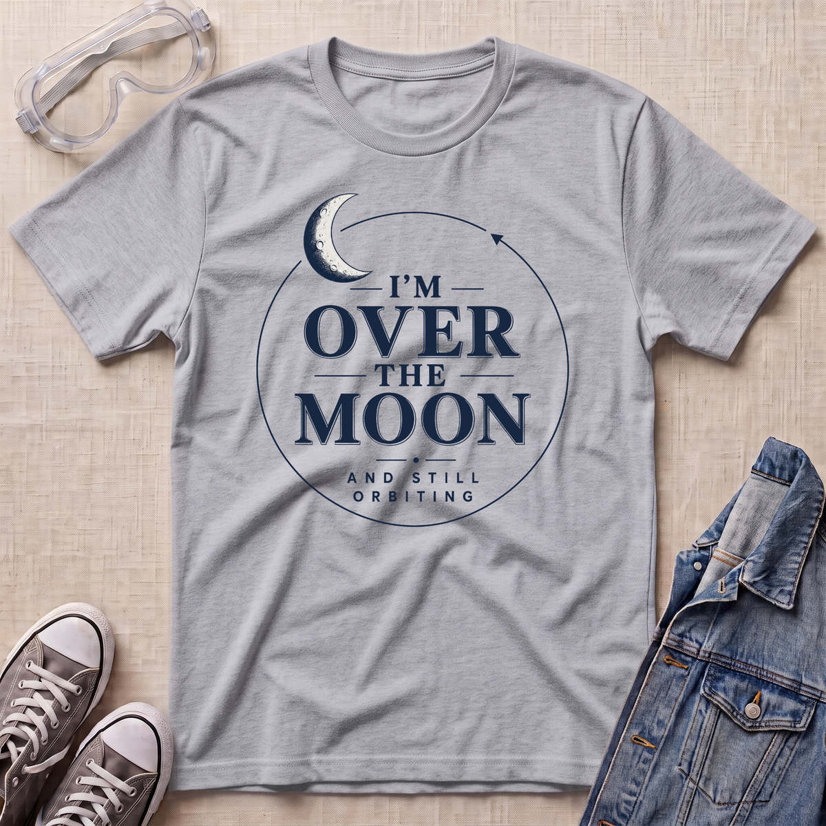 A sport grey graphic t-shirt laid flat on a beige textured background, styled with safety glasses, gray sneakers, and a denim jacket. The t-shirt features a navy blue graphic of a crescent moon inside a circle with the text 'I'M OVER THE MOON AND STILL ORBITING'.